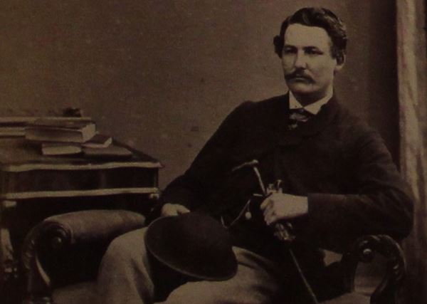 photo of a man sitting casually with a stick and bowler hat on his lap and a pile of books on a small desk beside him.